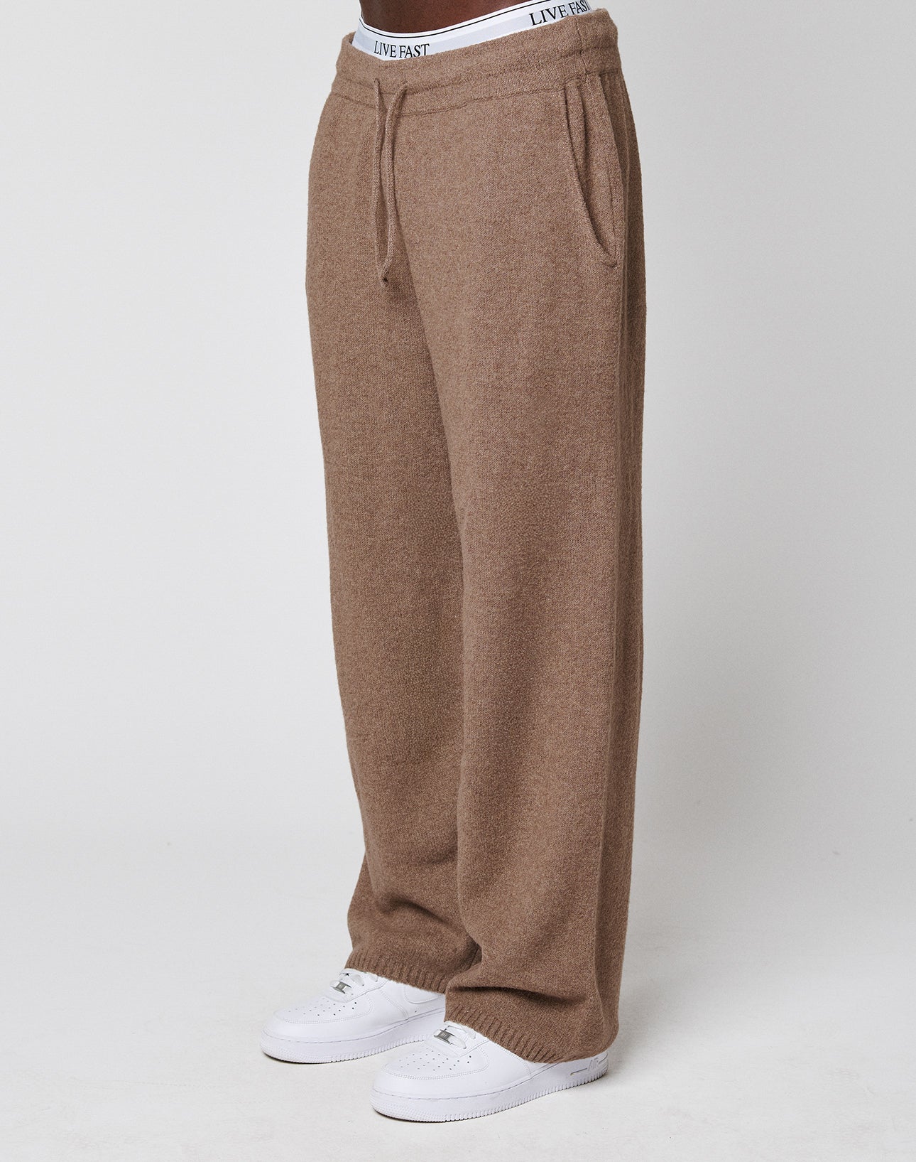 A person wears the LIVE FAST DIE YOUNG Live Fast Boucle Pants in loose-fitting brown knit with an elastic waistband and straight leg, paired with white sneakers and a visible "LIVE FAST" waistband, against a plain white background. | model