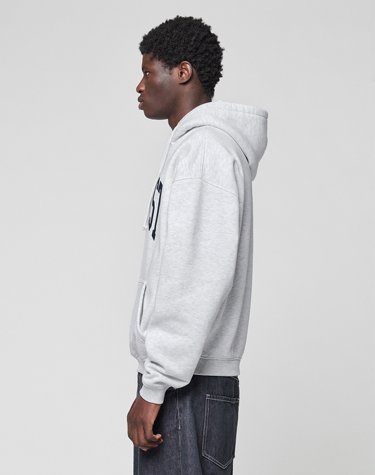 A person with short curly hair stands in profile, wearing a LIVE FAST DIE YOUNG Live Fast College Hooded sweatshirt in light gray with front pocket and dark jeans, set against a plain light gray background.