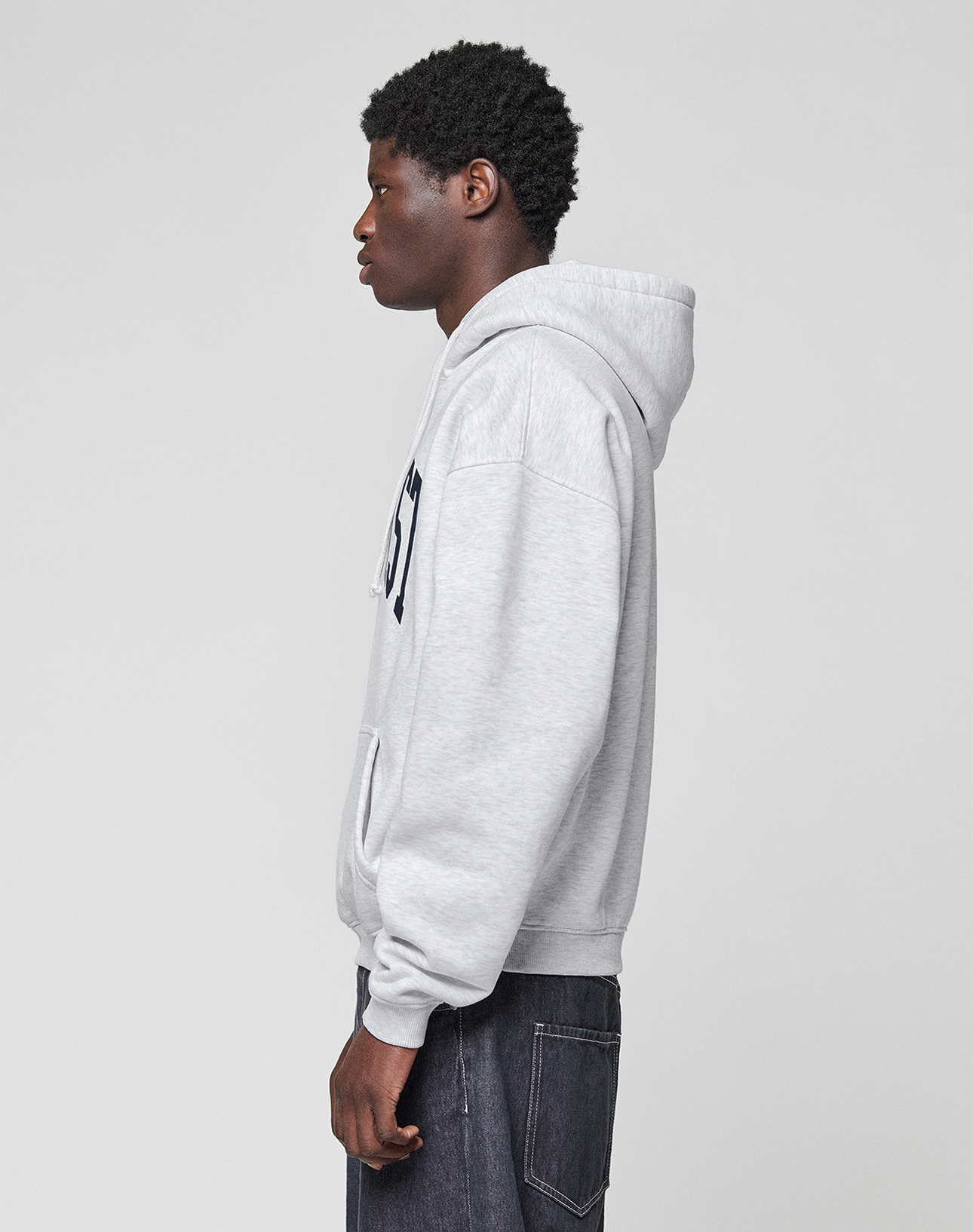 A person with short curly hair stands in profile, wearing a LIVE FAST DIE YOUNG Live Fast College Hooded sweatshirt in light gray with front pocket and dark jeans, set against a plain light gray background.