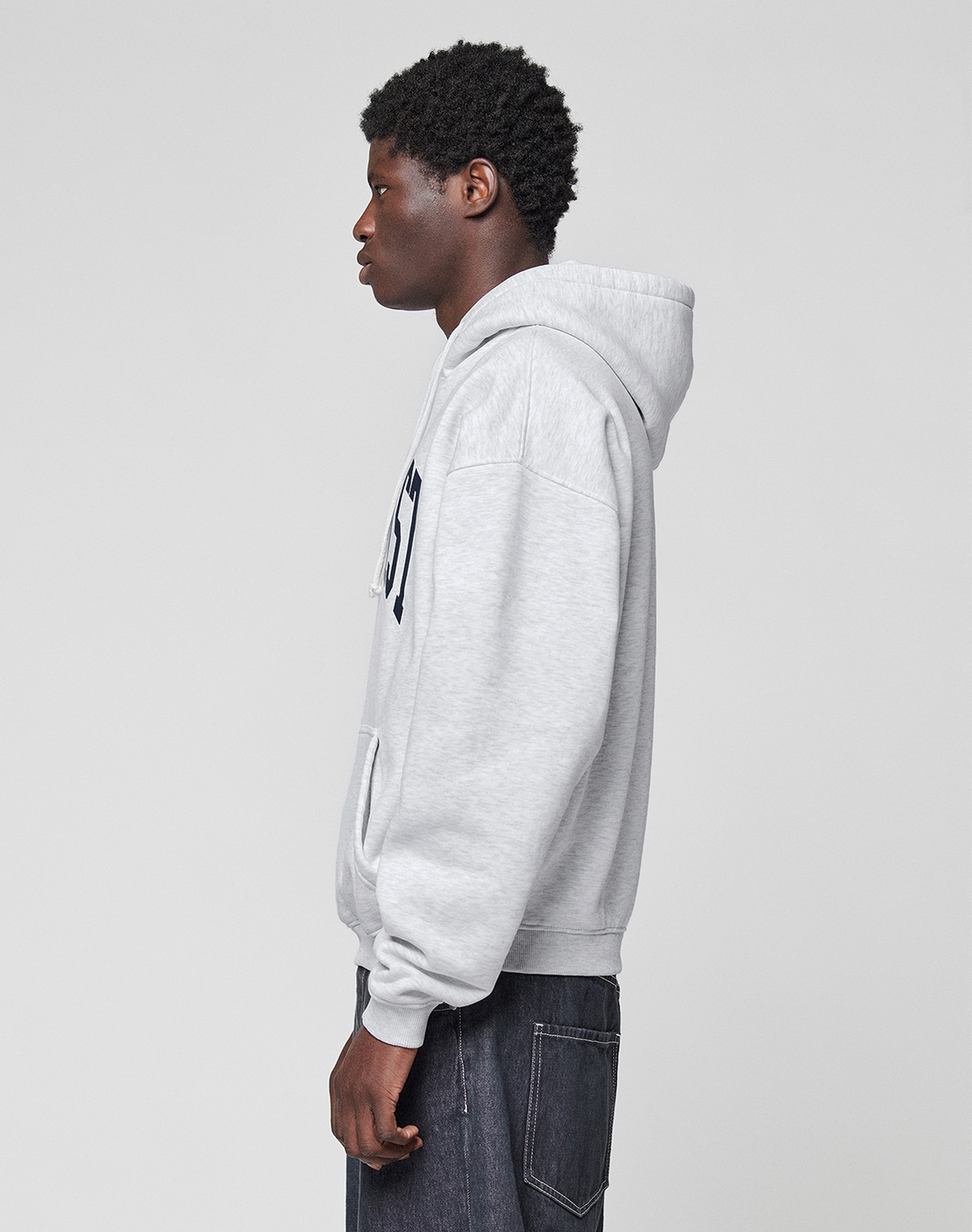 A person with short curly hair stands in profile, wearing a LIVE FAST DIE YOUNG Live Fast College Hooded sweatshirt in light gray with front pocket and dark jeans, set against a plain light gray background.