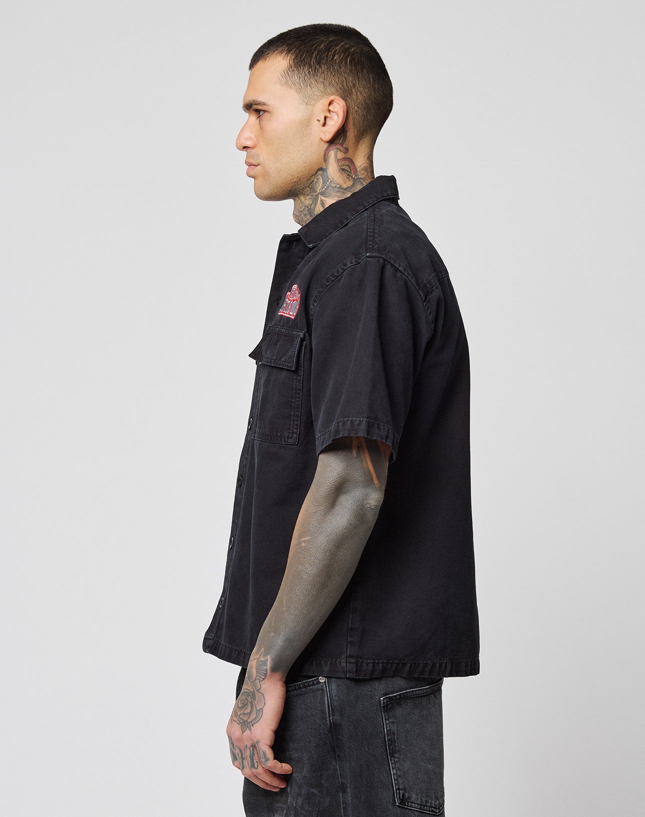 A tattooed man in profile wears the LFDY Workwear Shirt by LIVE FAST DIE YOUNG—an oversize black Baumwolle button-up with chest pockets—paired with dark jeans, set against a plain light background.