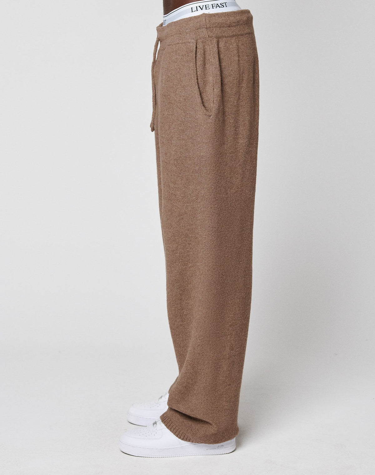 A person in LIVE FAST DIE YOUNG’s Live Fast Boucle Pants—brown, wide fit, loose knit, with a straight leg and elastic waistband featuring a “LIVE FAST” logo—paired with white sneakers, shown in side profile against a light background.