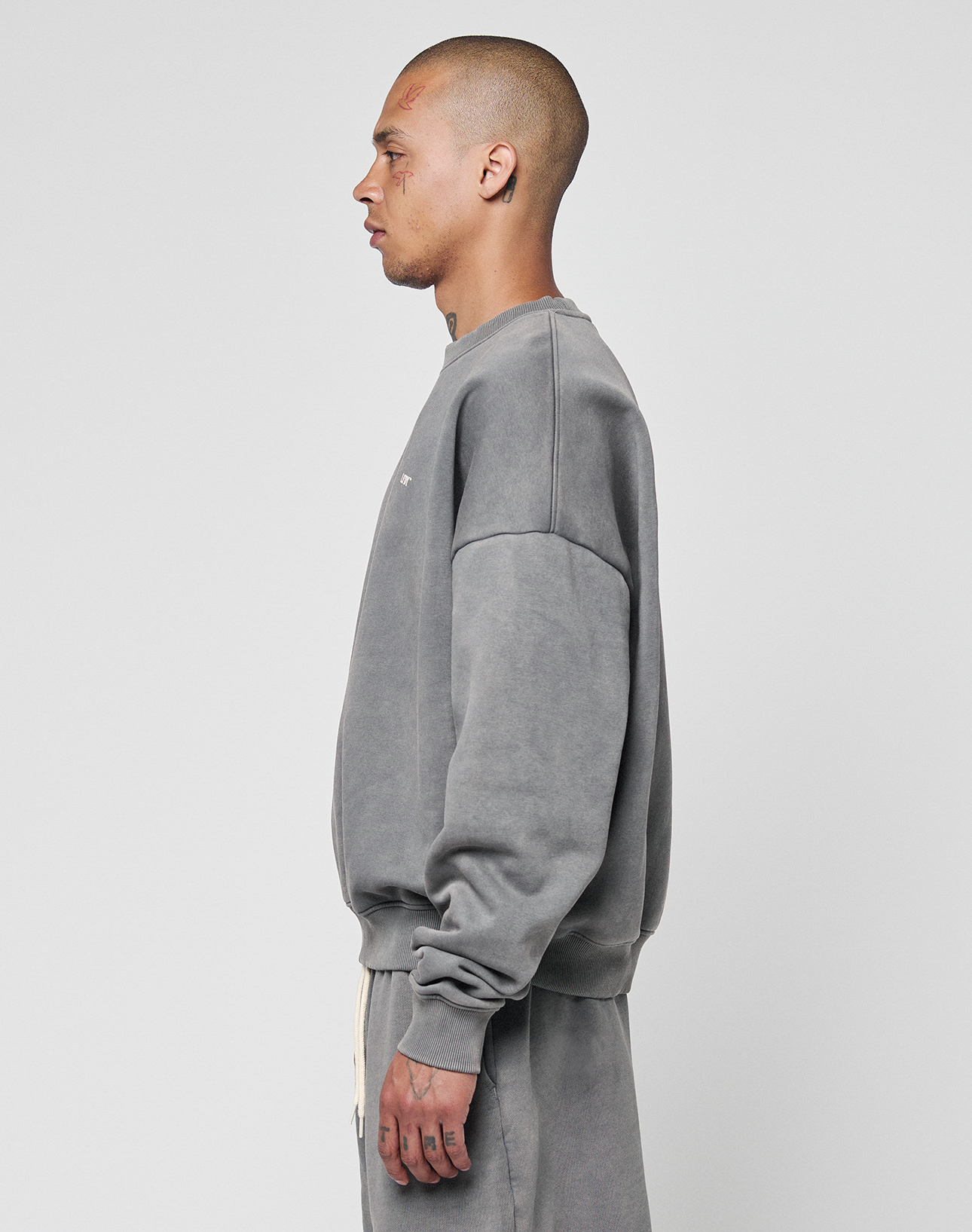 A person with a shaved head is shown in profile, wearing the LIVE FAST DIE YOUNG Basic Chest Sweater in gray and matching pants, styled in an oversize fit, against a plain white background.