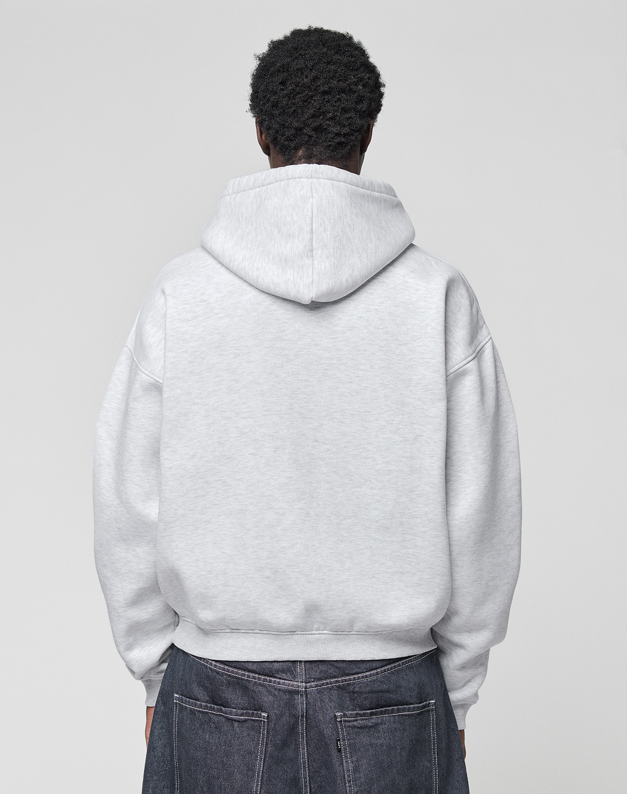 A person faces away from the camera, wearing a LIVE FAST DIE YOUNG Live Fast College Hooded oversize light gray sweatshirt and dark denim jeans against a plain light background.
