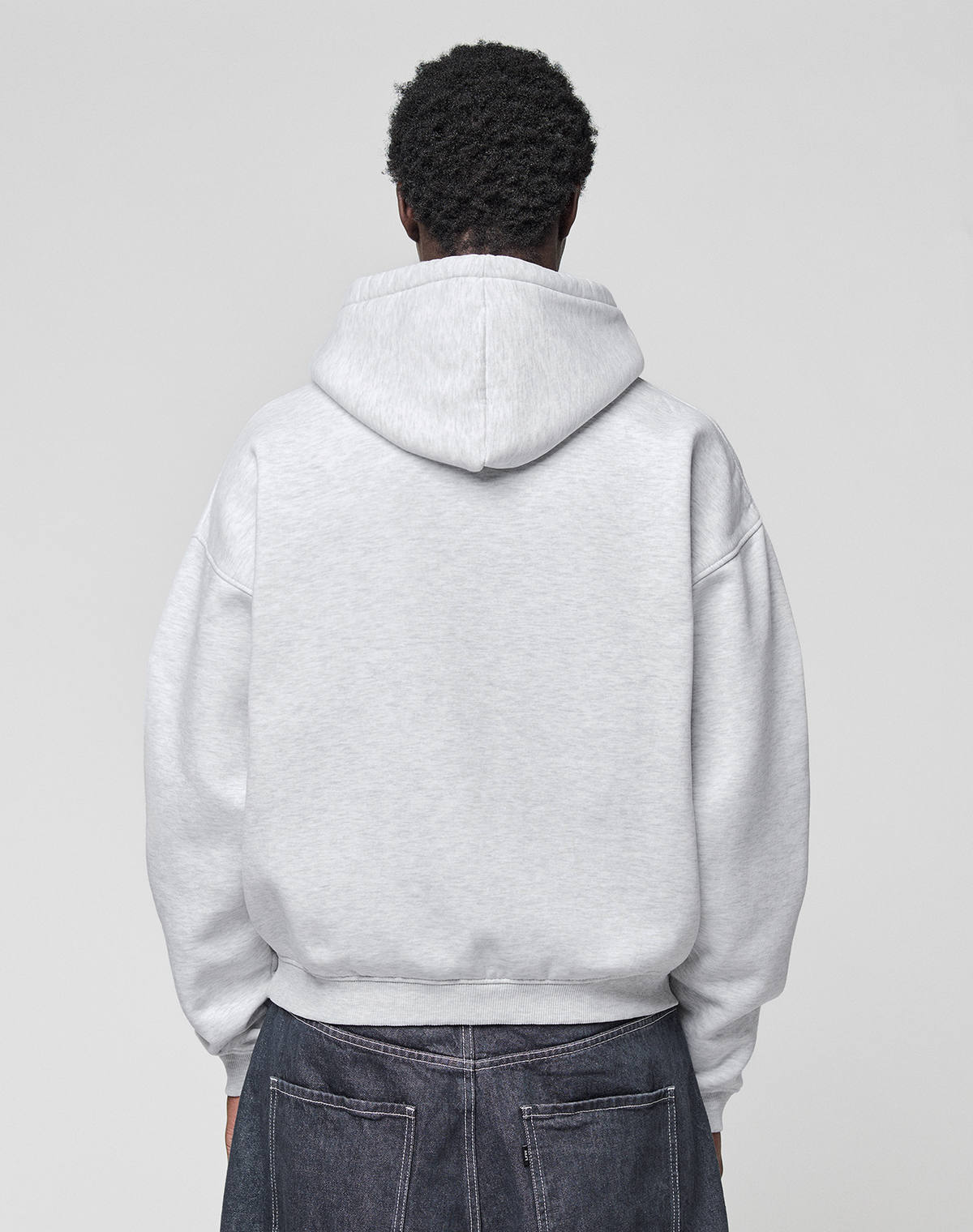 A person faces away from the camera, wearing a LIVE FAST DIE YOUNG Live Fast College Hooded oversize light gray sweatshirt and dark denim jeans against a plain light background.
