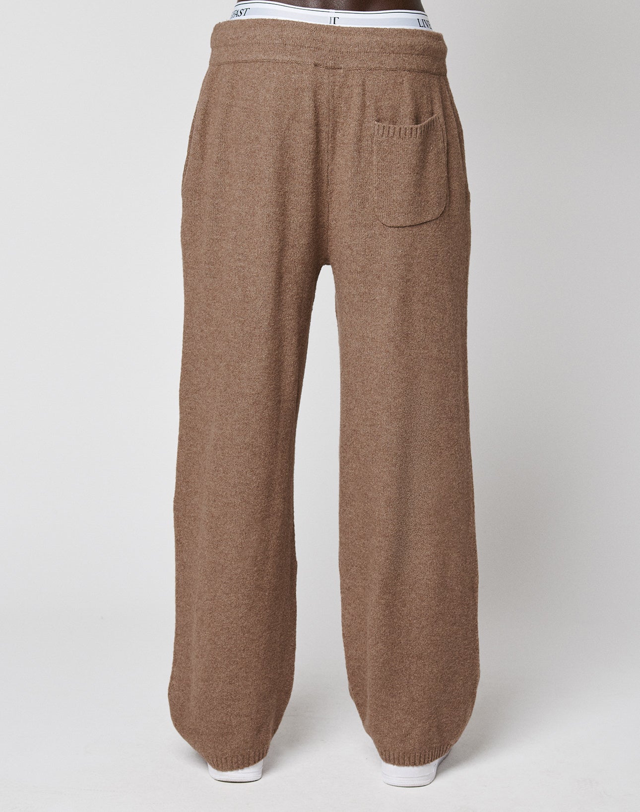 A person wearing LIVE FAST DIE YOUNG's Live Fast Boucle Pants—loose-fitting brown knit with an elastic waistband, straight leg, and a single back pocket—stands against a plain light background, with white underwear visible above the pants.