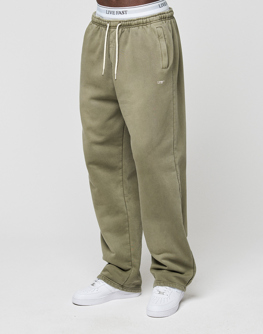 A person wears the LIVE FAST DIE YOUNG Basic Wide Jogger in loose olive green with a white drawstring, featuring a white logo-print waistband and white sneakers, standing against a plain white background.