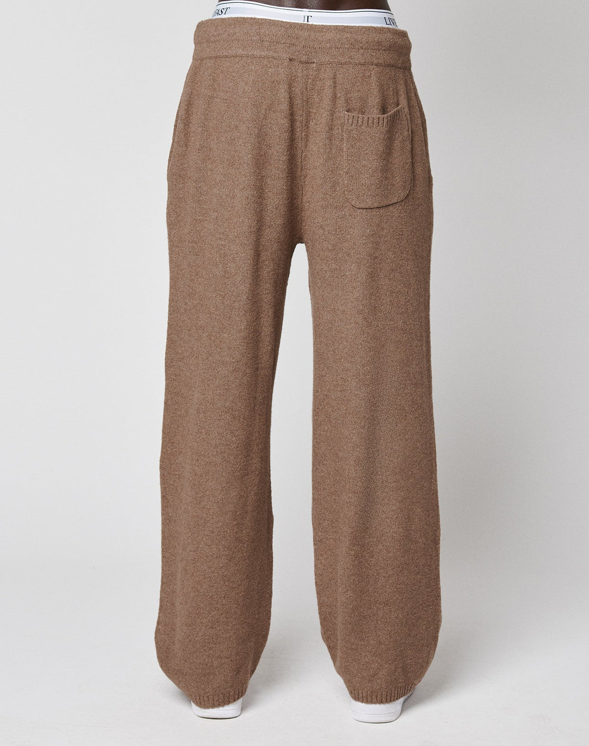 A person wearing LIVE FAST DIE YOUNG's Live Fast Boucle Pants—loose-fitting brown knit with an elastic waistband, straight leg, and a single back pocket—stands against a plain light background, with white underwear visible above the pants.