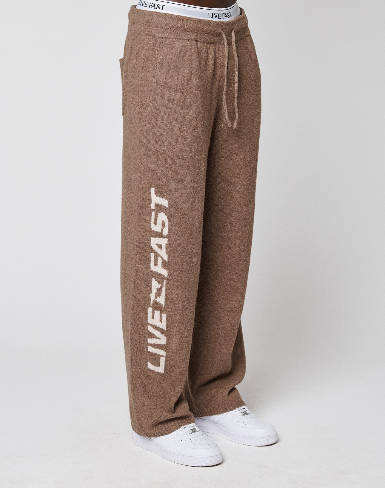 A person wears LIVE FAST DIE YOUNG's Live Fast Boucle Pants, wide fit and brown with white "LIVE FAST" on the left leg and elastic waistband. Styled with white sneakers against a plain light background.