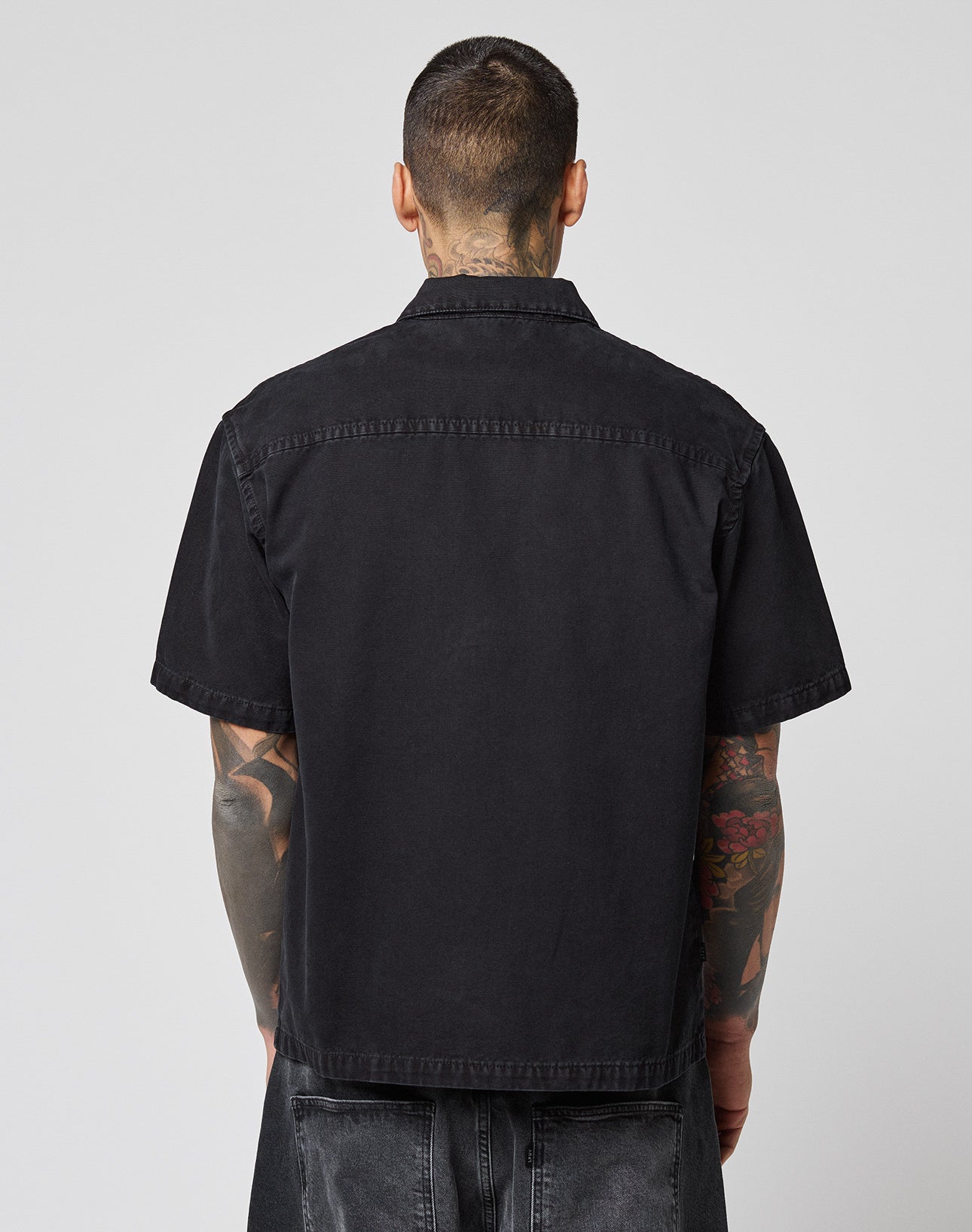 A person with short hair and visible arm tattoos is shown from behind, wearing the black LFDY Workwear Shirt by LIVE FAST DIE YOUNG and dark jeans, standing against a plain gray background.