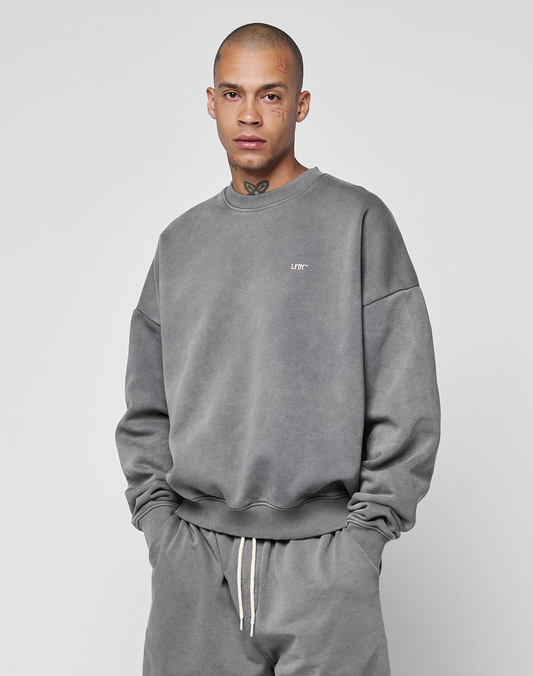 A tattooed person with a shaved head wears the LIVE FAST DIE YOUNG Basic Chest Sweater in gray and matching drawstring pants, standing against a plain light gray background.