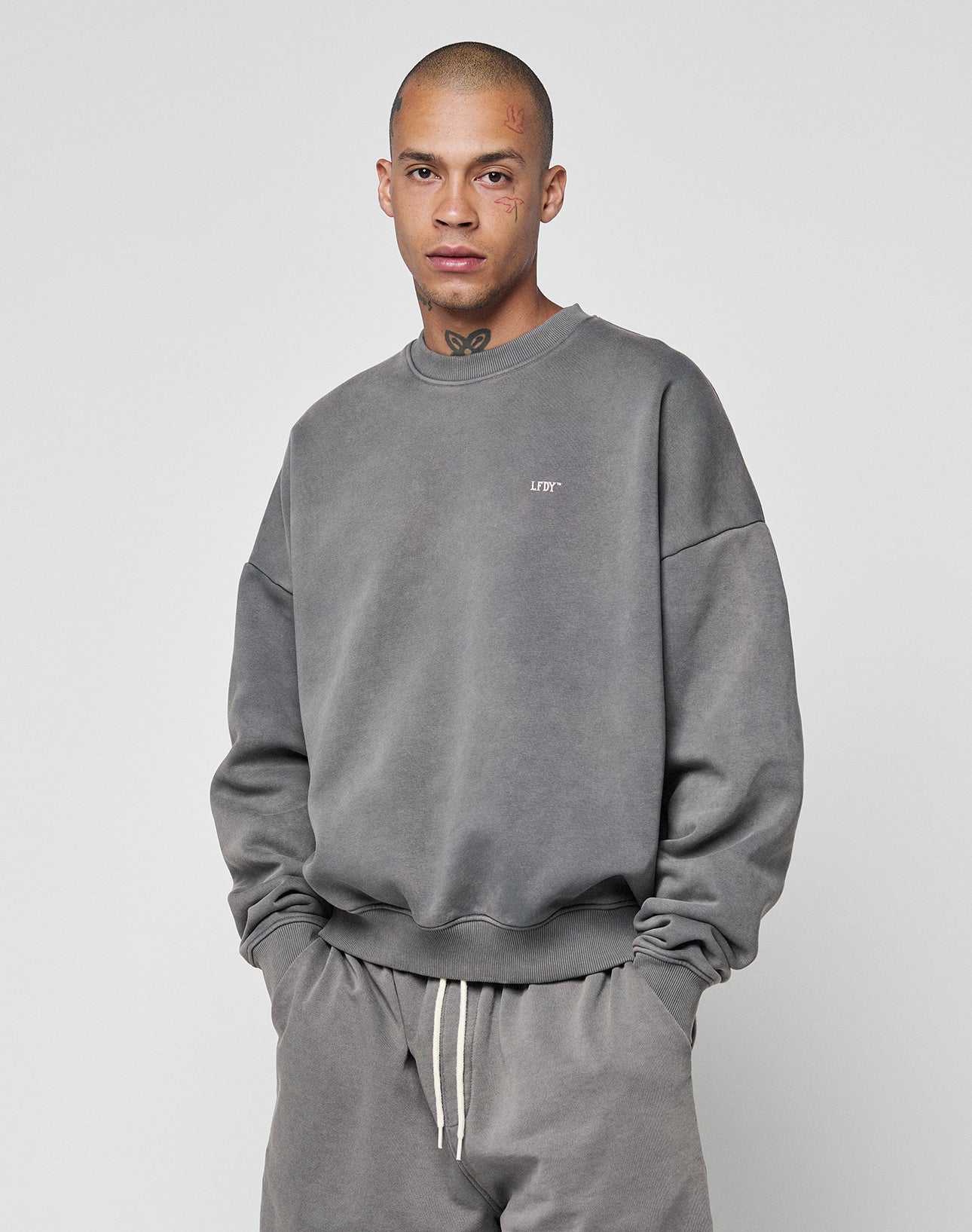 A person with a shaved head and face/neck tattoos wears the LIVE FAST DIE YOUNG Basic Chest Sweater in an oversized, faded gray style with matching sweatpants, hands in pockets, standing against a plain background.