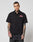 A tattooed man wears the LIVE FAST DIE YOUNG LFDY Workwear Shirt, an oversize black cotton short-sleeve button-up with two chest pockets and a red embroidered patch, standing against a plain light background.