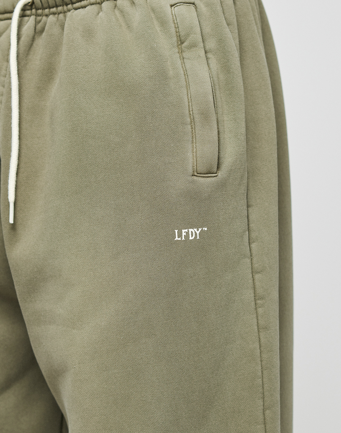 Close-up of the LIVE FAST DIE YOUNG Basic Wide Jogger in olive green, featuring a wide fit, elastic waistband, cream drawstring, slanted pocket, and a small white LFDY logo embroidered on the upper thigh.