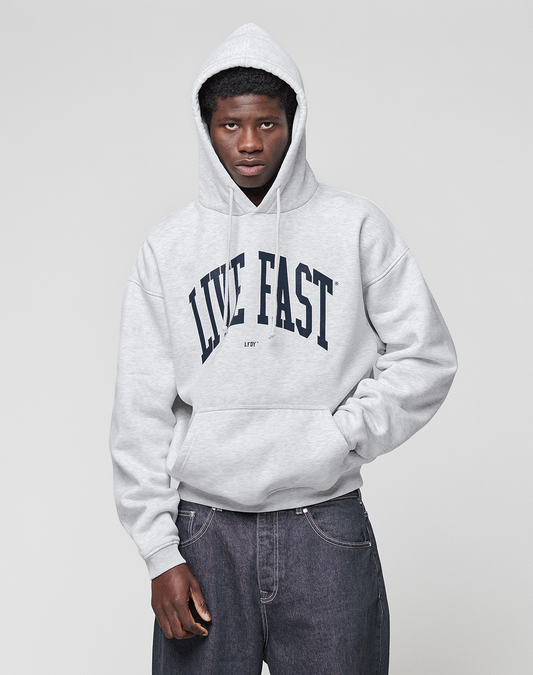 A person wears the LIVE FAST DIE YOUNG Live Fast College Hooded, an oversized light gray cotton hoodie with a "LIVE FAST" front print, standing against a plain gray background with hands in pockets and a neutral expression.