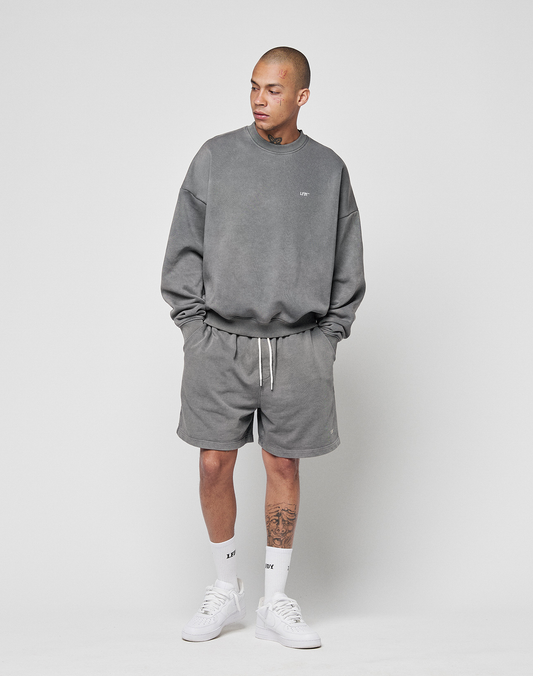 A person stands against a plain background, wearing the LIVE FAST DIE YOUNG Basic Chest Sweater in gray with an oversize fit and matching gray shorts, white crew socks, and white sneakers. They have a tattoo on their left leg and are looking to the side.