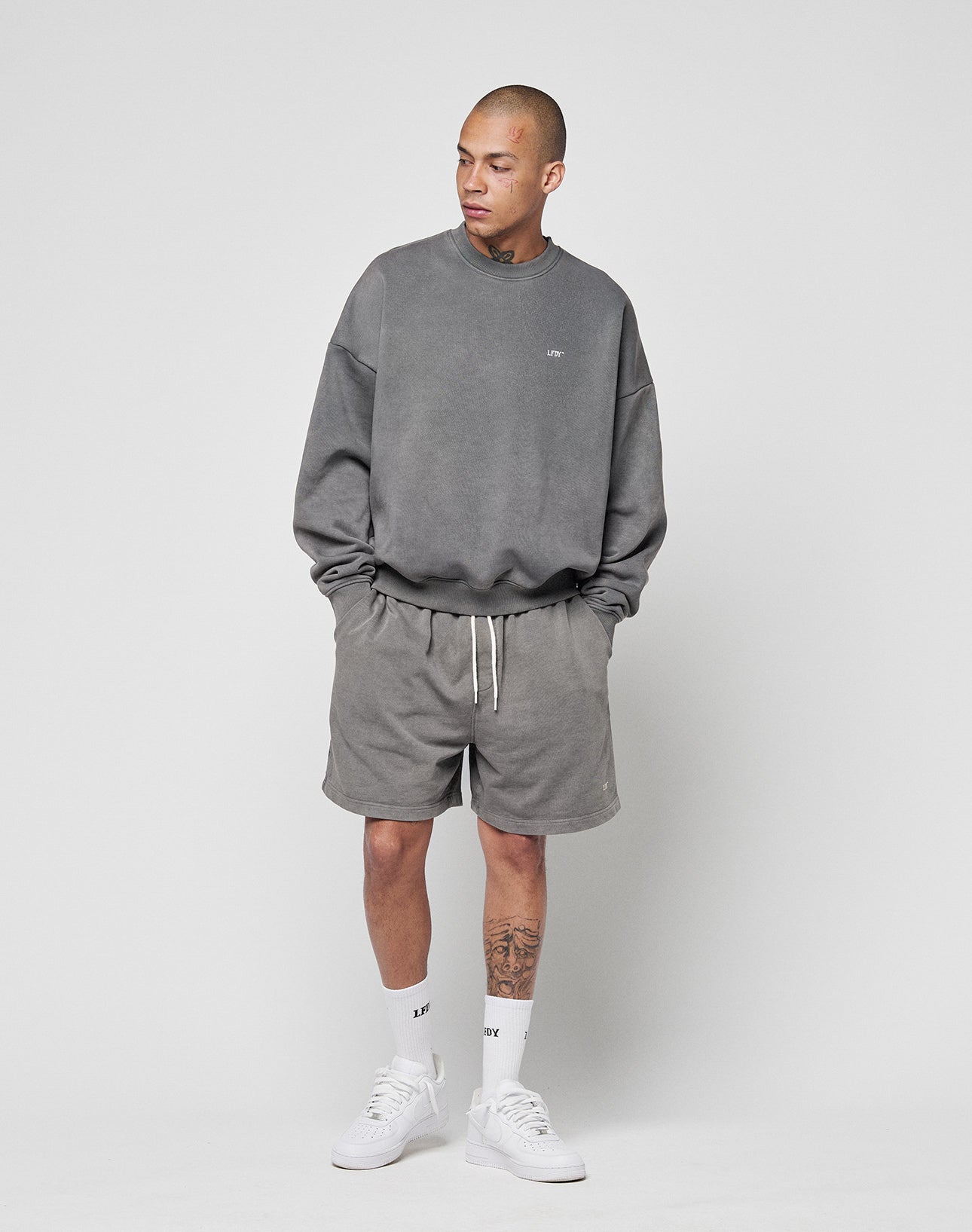 A person stands against a plain background wearing the LIVE FAST DIE YOUNG Basic Chest Sweater in grey, matching grey shorts, white crew socks, and white sneakers. They have a tattoo on their left leg and are looking to the side.