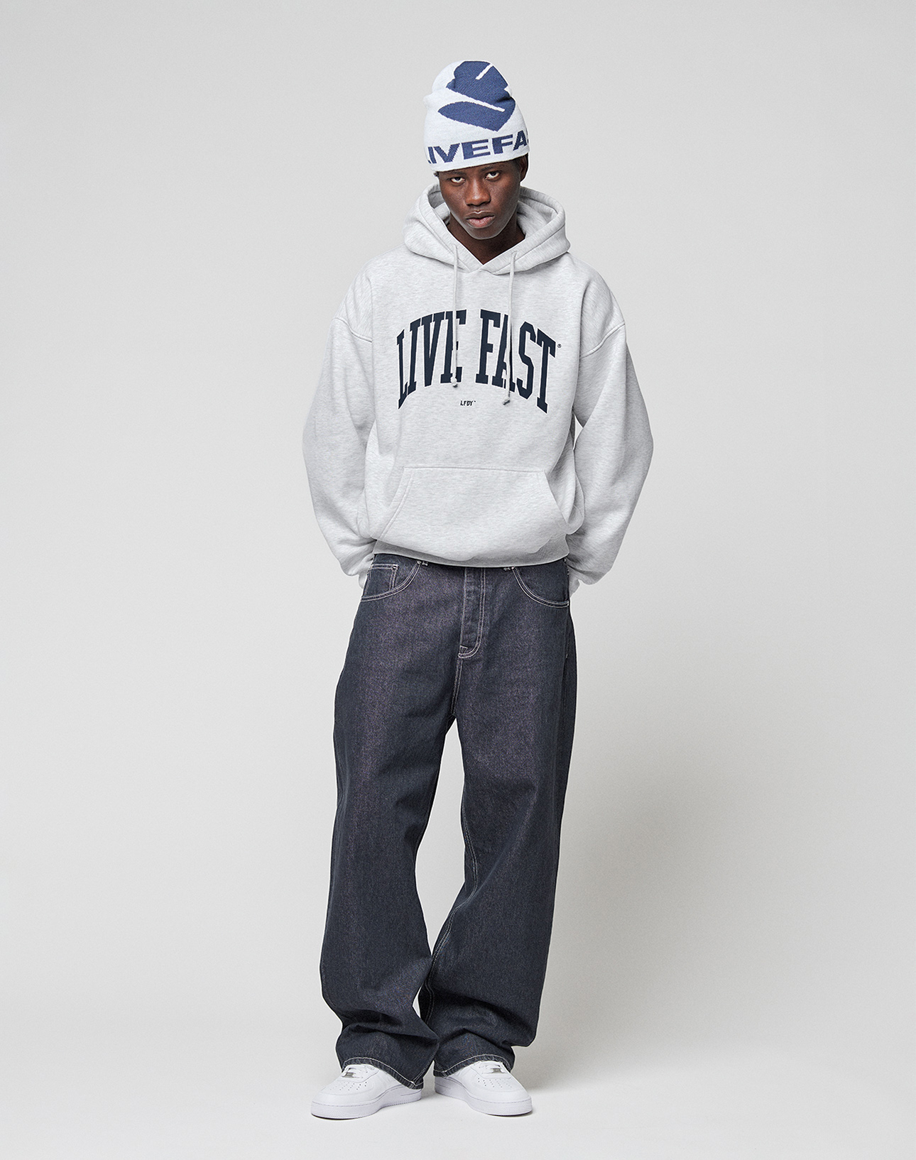 A person stands against a plain background wearing a gray LIVE FAST DIE YOUNG "Live Fast College Hooded" with an oversized fit and bold front print, styled with dark baggy jeans, white sneakers, and a white beanie featuring blue graphics. Hands in pockets, relaxed pose.