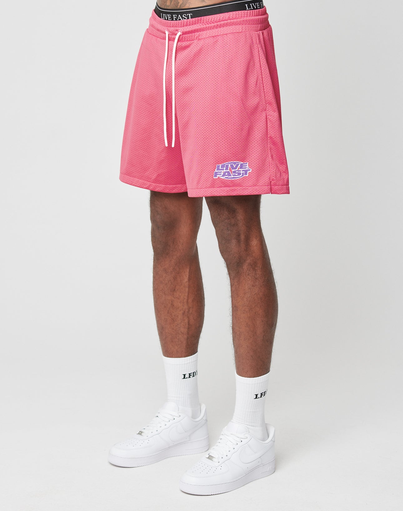 A person wears LFDY Oval Mesh Shorts by LIVE FAST DIE YOUNG with an elastic waistband and regular fit, plus white crew socks—both featuring LIVE FAST text—and white Nike sneakers, standing against a plain white background. | model