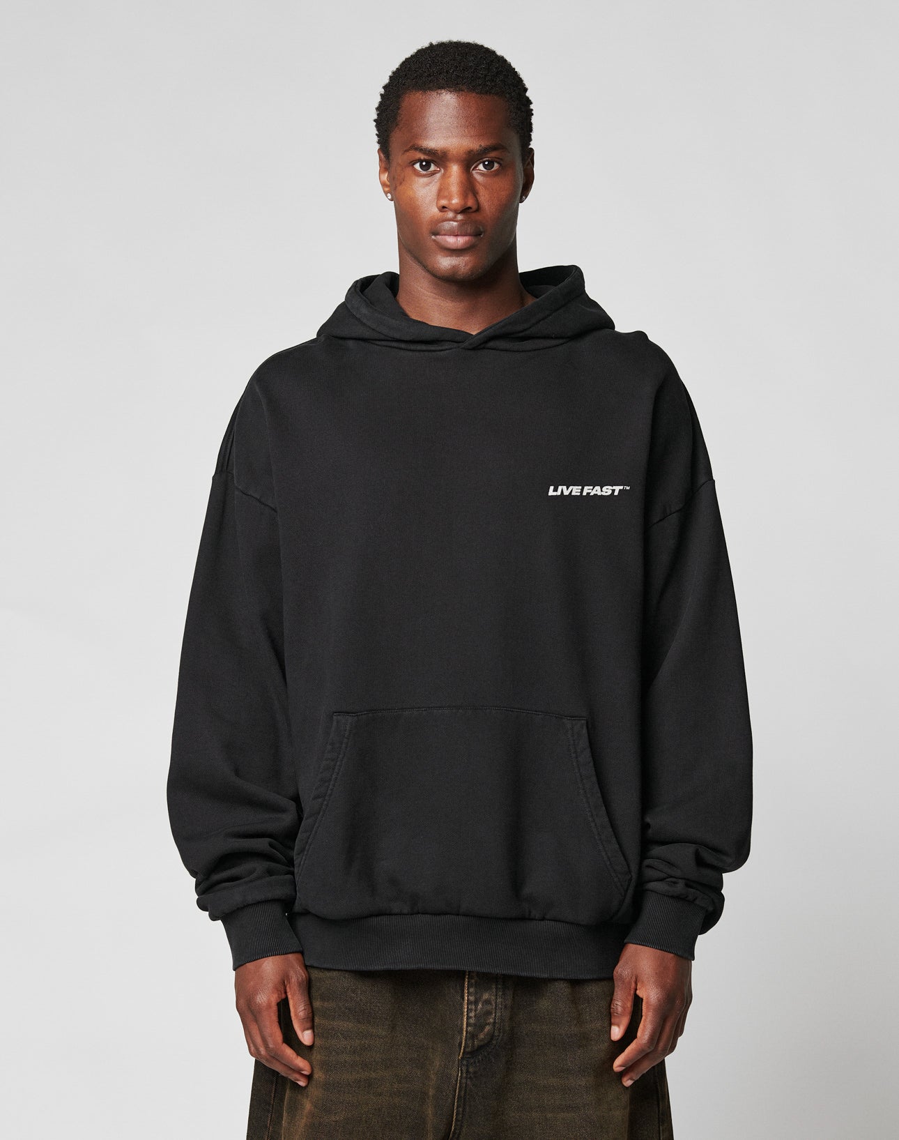 A person faces forward in an oversize fit LFDY Hardwear Hooded black hoodie by LIVE FAST DIE YOUNG, made from Baumwolle with LIVE FAST in small white letters on the left chest, paired with dark pants against a plain light background.