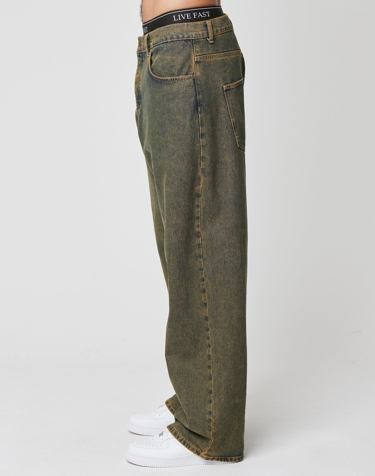 A person stands in profile wearing LFDY Baggy Denim by LIVE FAST DIE YOUNG—loose-fitting, faded olive green jeans with baggy fit, wide straight leg, yellow stitching, back pockets, and a LIVE FAST waistband label. 100% Baumwolle. White background.