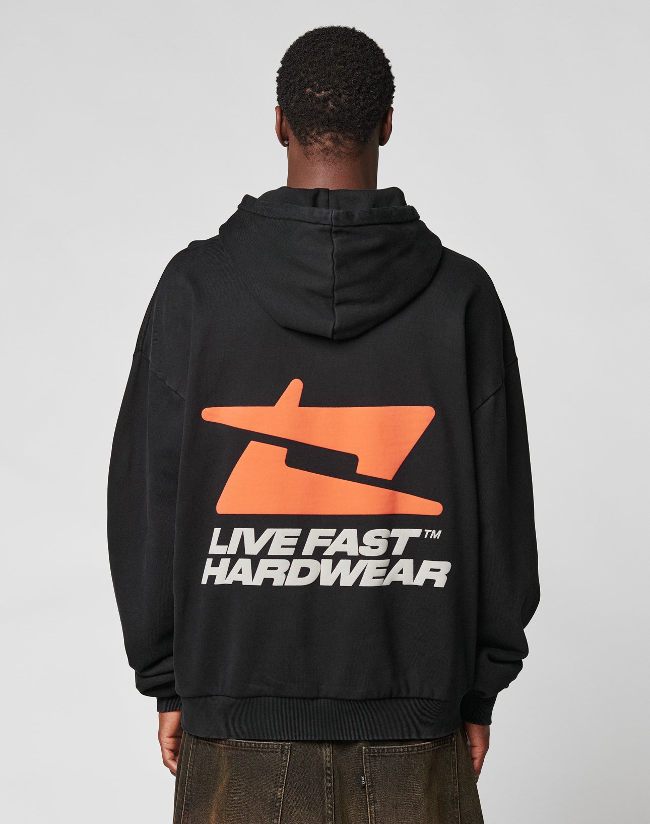 A person in a black LFDY Hardwear Hooded by LIVE FAST DIE YOUNG, made from Baumwolle with an orange abstract graphic and LIVE FAST HARDWEAR on the back, stands against a plain light gray background.