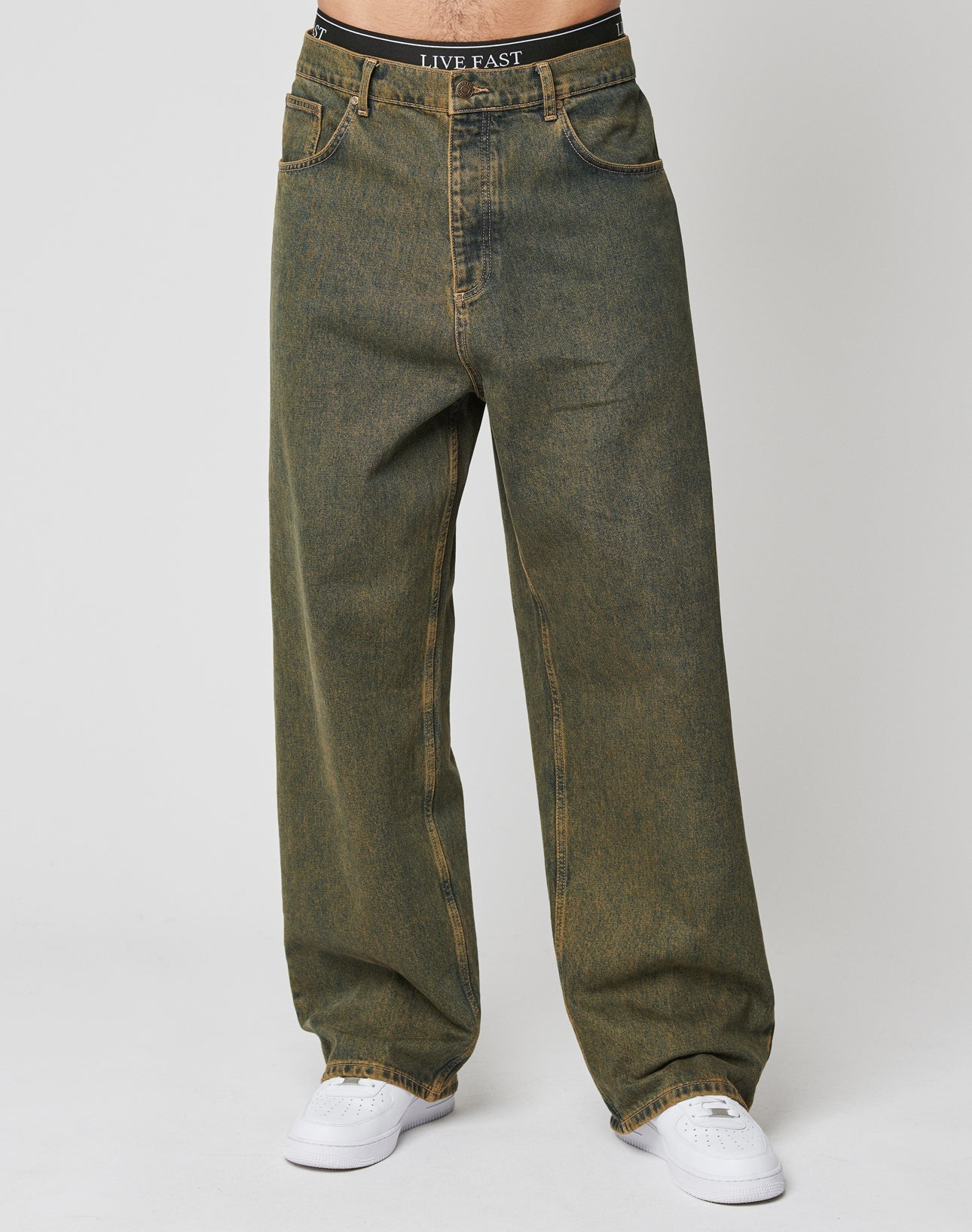 A model wears LFDY Baggy Denim by LIVE FAST DIE YOUNG—faded green, high-waisted, wide-leg, 100% cotton jeans with a baggy fit—paired with white sneakers and a black LIVE FAST waistband, set against a plain white background.