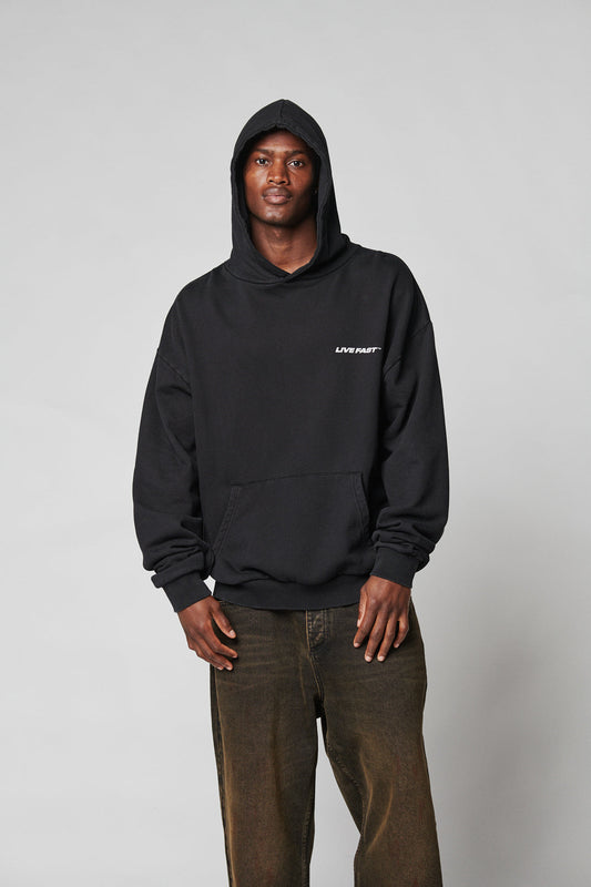 A person wears a black LFDY Hardwear Hooded by LIVE FAST DIE YOUNG with the hood up and chest logo, paired with loose-fitting faded brown Oversize Fit pants, standing against a plain light gray background. The hoodie is 100% Baumwolle.
