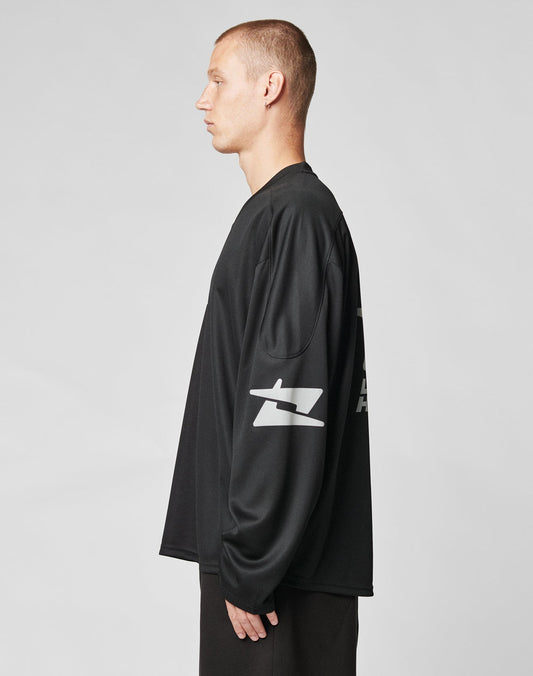 A person with short hair stands in profile, facing left, wearing the oversize fit Live Fast Jersey by LIVE FAST DIE YOUNG, featuring a reflective print on the sleeve. The background is plain light gray.