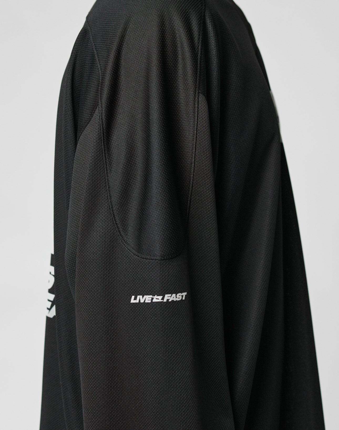 Close-up of the Live Fast Jersey by LIVE FAST DIE YOUNG—an oversized black long-sleeve shirt with LIVE FAST in a white reflective print on the upper sleeve. Made of breathable polyester athletic fabric. Plain, light background.