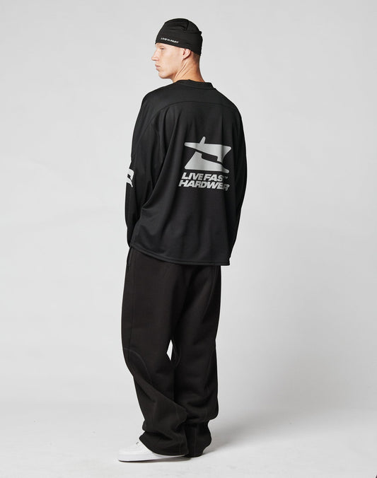A person stands sideways in a black beanie, loose black pants, white sneakers, and the LIVE FAST DIE YOUNG Live Fast Jersey—a polyester oversize long-sleeve with a shark graphic and LIVE FAST HARDWEAR reflective print on the back—against a plain light background.