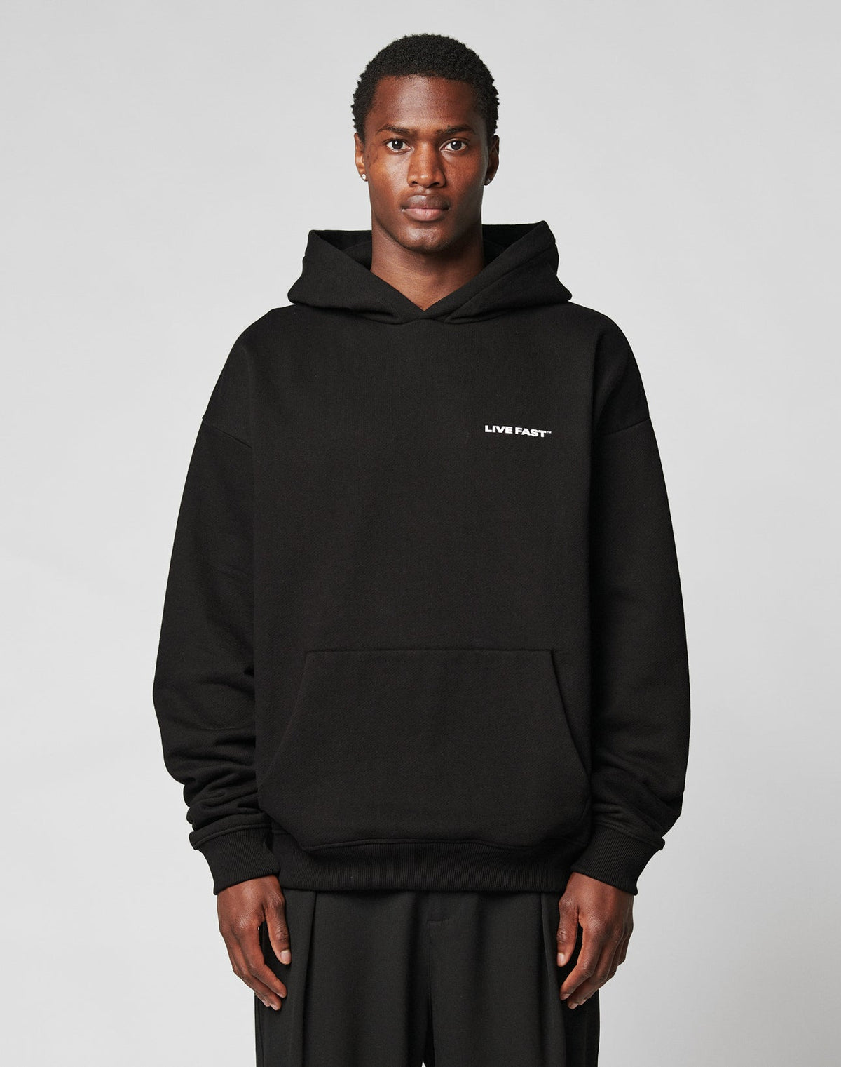 A person in the Live Fast Plain Hooded by LIVE FAST DIE YOUNG—a black, oversized cotton-polyester hoodie with LIVE FAST in white on the left chest—stands against a plain light gray background. | model