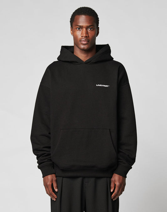 A person in the Live Fast Plain Hooded by LIVE FAST DIE YOUNG—a black, oversized cotton-polyester hoodie with LIVE FAST in white on the left chest—stands against a plain light gray background. | model