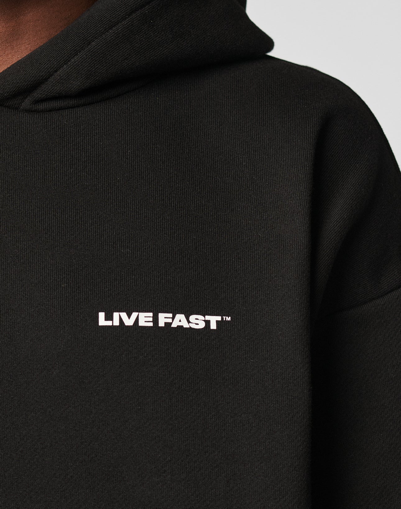 Close-up of the black Live Fast Plain Hooded by LIVE FAST DIE YOUNG, featuring an oversize fit and LIVE FAST™ in white uppercase letters on the chest. Made from a soft cotton-polyester blend; only part of the shoulder, neck, and hood are visible.