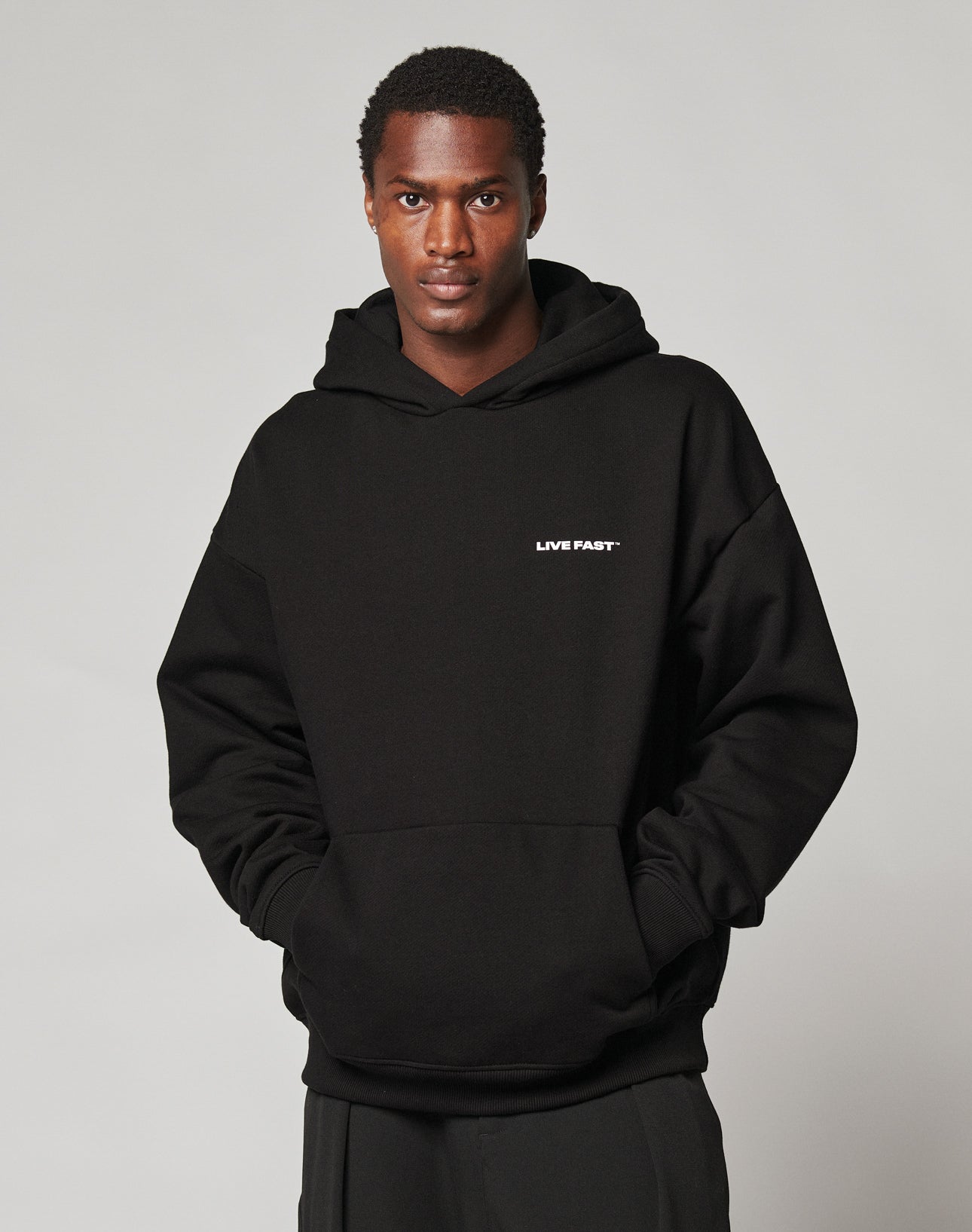 A person wears the LIVE FAST Plain Hooded hoodie by LIVE FAST DIE YOUNG, featuring an oversize black fit with LIVE FAST in small white letters on the chest, hands in pockets. Crafted from a soft cotton-polyester blend against a plain light background.