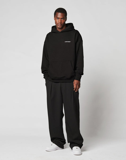 A person stands against a plain light background, wearing a black hoodie with small white text on the left chest, LIVE FAST DIE YOUNG Wide Casual Pants, and white sneakers. They look directly at the camera with a neutral expression.