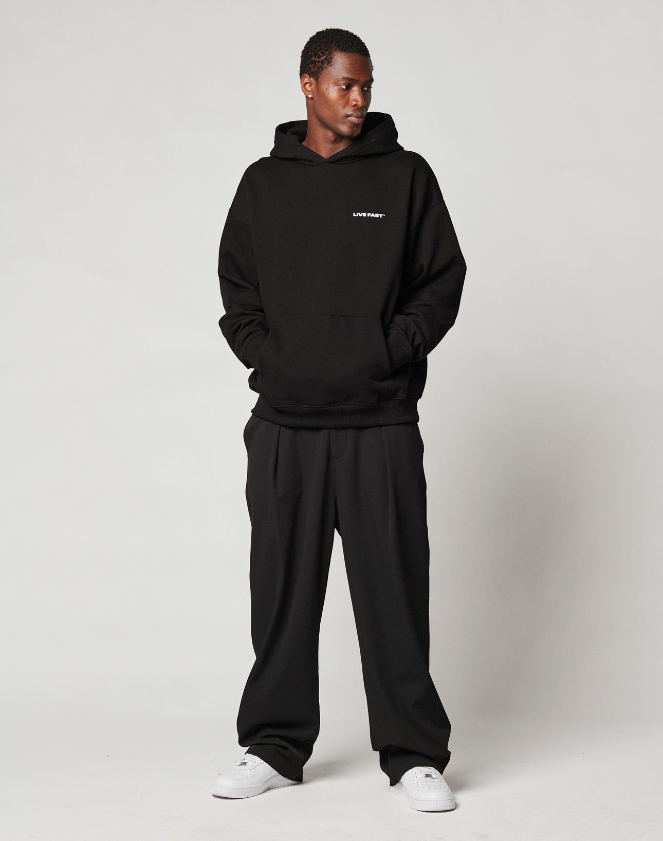 A person stands against a plain light background wearing a black LIVE FAST DIE YOUNG hoodie, Wide Casual Pants with an elastic waistband and side pockets, and white sneakers, looking to the side with hands in the hoodie pocket.