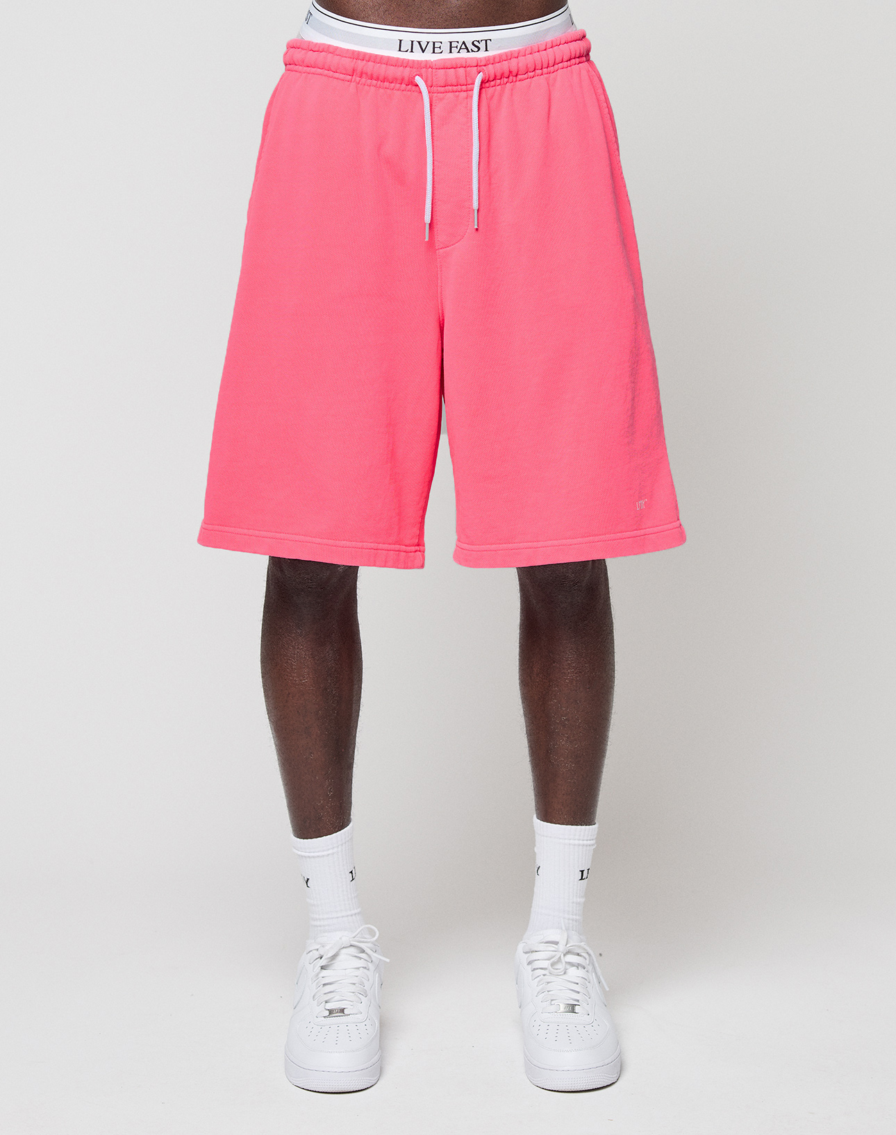 A person in bright pink Basic Shorts by LIVE FAST DIE YOUNG, with an elastic waistband, white socks, and white sneakers stands against a plain white background. The waistband displays the LIVE FAST branding on their underwear.