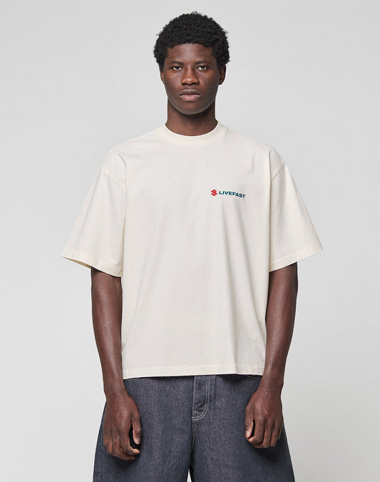 A person stands against a plain background wearing the LIVE FAST DIE YOUNG LFDY Dealership Tee—an oversize, off-white 100% Baumwolle T-shirt with a small red and blue LIVEFAST logo on the left chest—paired with dark wide-leg jeans.