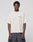 A person stands against a plain background wearing the LIVE FAST DIE YOUNG LFDY Dealership Tee—an oversize, off-white 100% Baumwolle T-shirt with a small red and blue LIVEFAST logo on the left chest—paired with dark wide-leg jeans.
