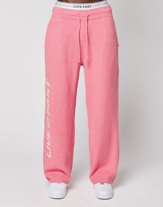 Someone is wearing the Live Fast Boucle Pants by LIVE FAST DIE YOUNG—pink sweatpants with "LIVE FAST" in white on the left leg, a comfy elastic waistband, and paired with matching white sneakers.
