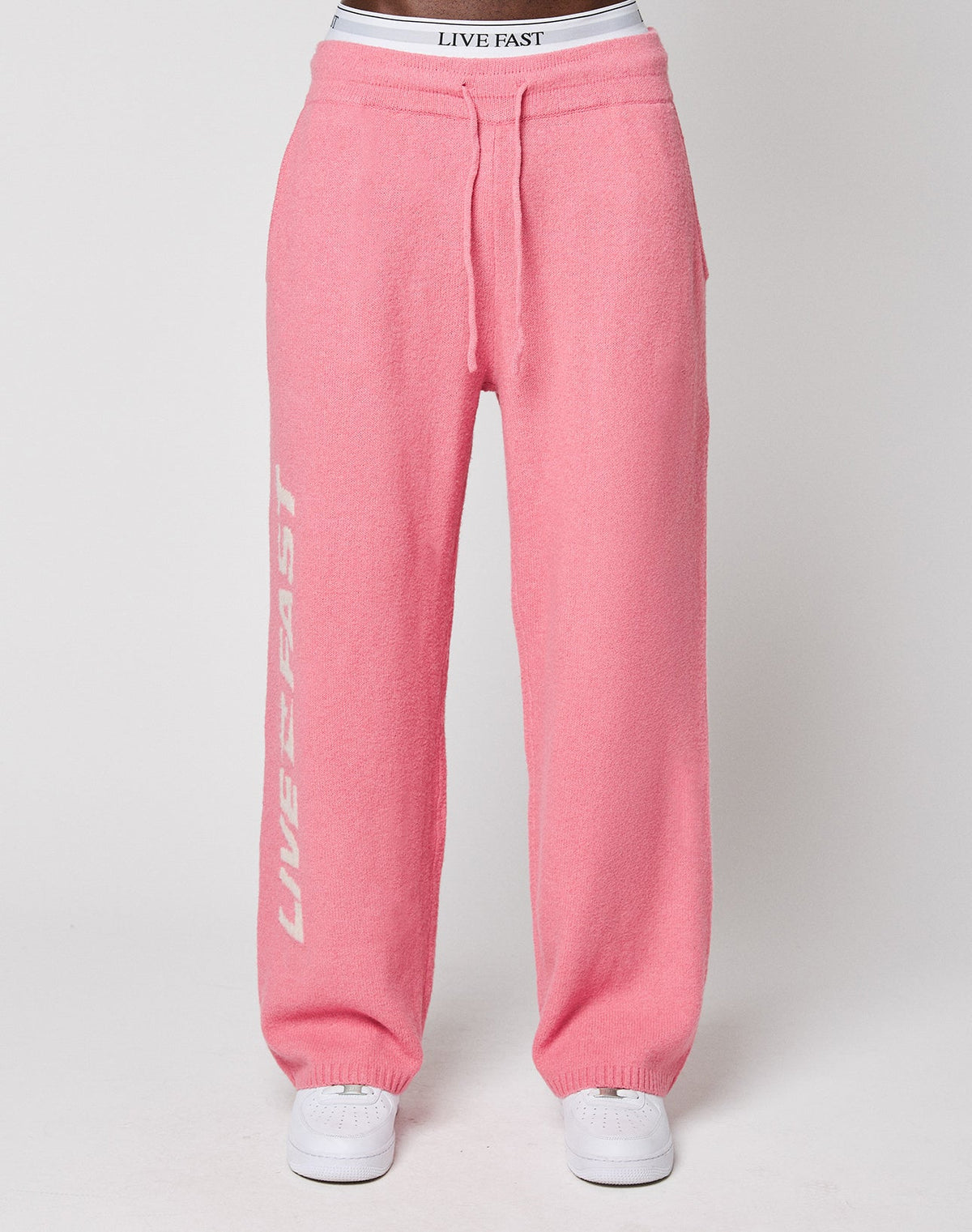 Someone is wearing the Live Fast Boucle Pants by LIVE FAST DIE YOUNG—pink sweatpants with "LIVE FAST" in white on the left leg, a comfy elastic waistband, and paired with matching white sneakers.