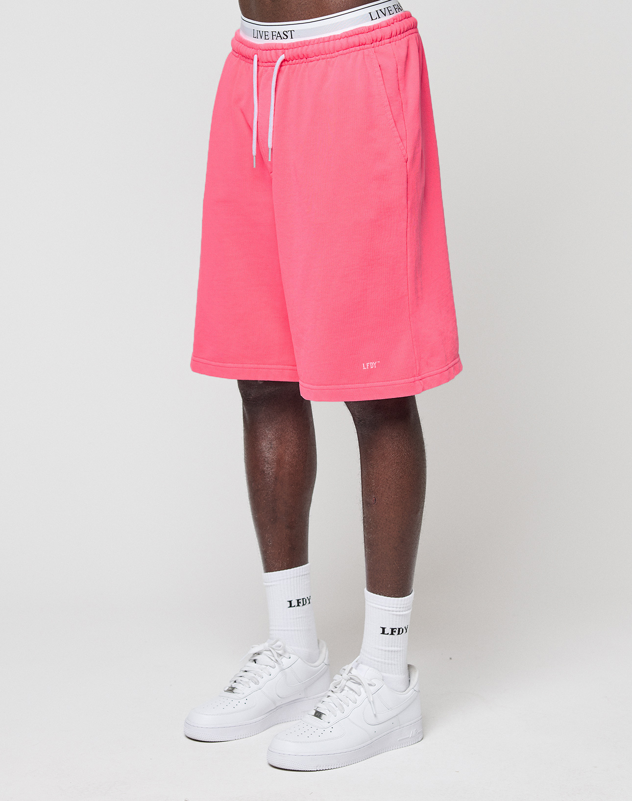 A person wears LIVE FAST DIE YOUNG Basic Shorts in bright pink with an elastic waistband, white socks with “LEDY” text, and white Nike sneakers. Only the lower half of their body is visible against a plain white background.