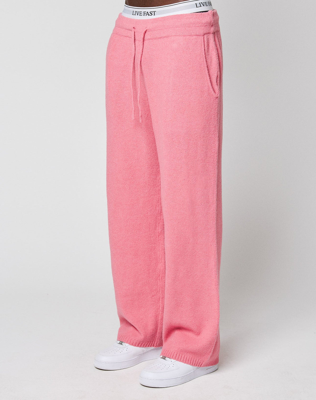 A person wears loose-fitting, wide fit pink Live Fast Boucle Pants by LIVE FAST DIE YOUNG, featuring an elastic waistband with "LIVE FAST" text. Styled with white sneakers against a plain, light background. | model