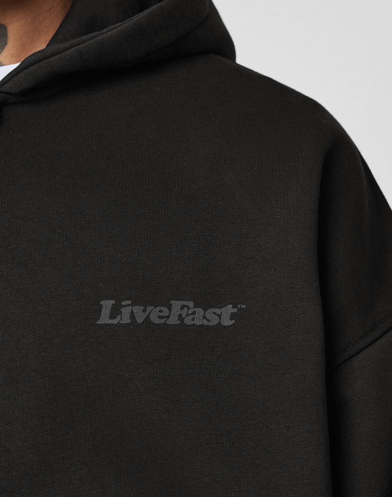 A close-up of a person wearing the LIVE FAST DIE YOUNG Basic 510 Hooded Zip in black with Vintage-Waschung and a subtle gray LiveFast chest print. The oversize fit highlights the neck tattoo of the wearer.