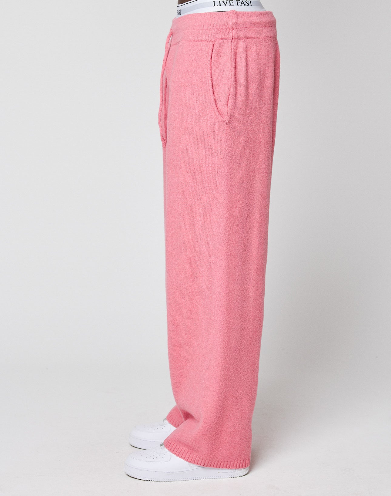 Wearing pink Live Fast Boucle Pants by LIVE FAST DIE YOUNG—loose fit, straight leg, elastic waistband with LIVE FAST logo, side pockets—paired with white sneakers on a plain white background.