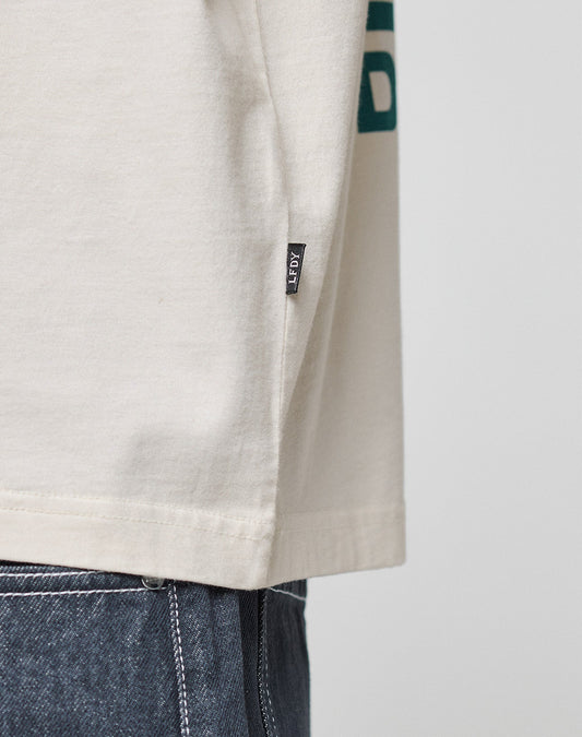 Close-up of the side of a beige LFDY Dealership Tee by LIVE FAST DIE YOUNG, made from 100% Baumwolle, featuring a small black ASRT tag near the hem. Paired with blue denim jeans against a plain, light background.