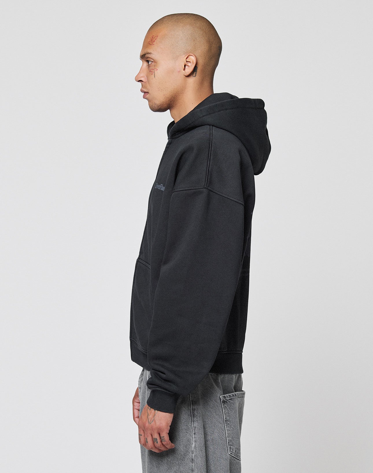 A person with a shaved head and face tattoos stands in profile wearing the LIVE FAST DIE YOUNG Basic 510 Hooded Zip in an oversize fit, paired with gray jeans, against a plain white background.