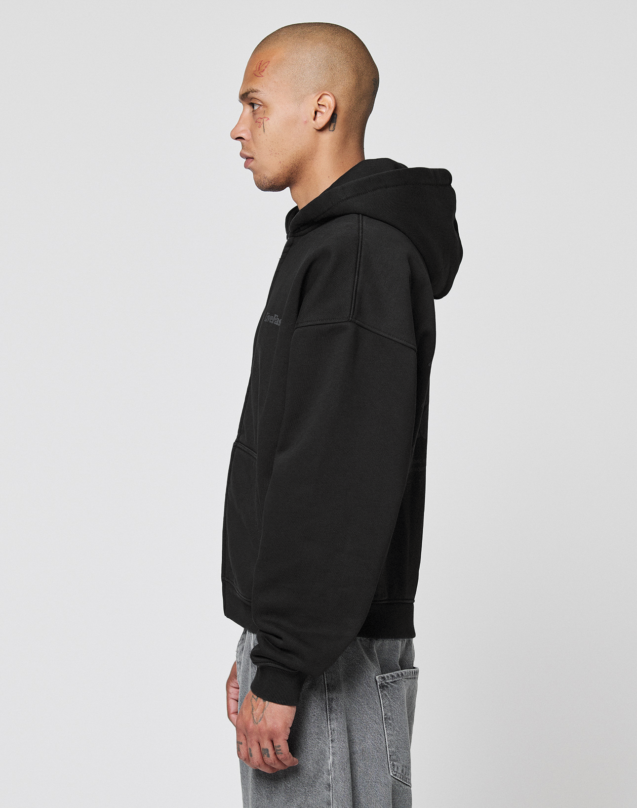 A person with a shaved head and face tattoos stands in profile wearing the LIVE FAST DIE YOUNG Basic 510 Hooded Zip in black and gray vintage-washed jeans against a plain light background.