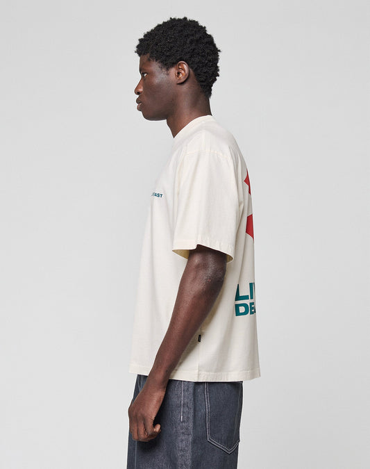 A person with short curly hair stands in profile against a plain background, wearing the LIVE FAST DIE YOUNG LFDY Dealership Tee—an oversize fit off-white T-shirt made from 100% Baumwolle with graphic front and back prints—and dark jeans.